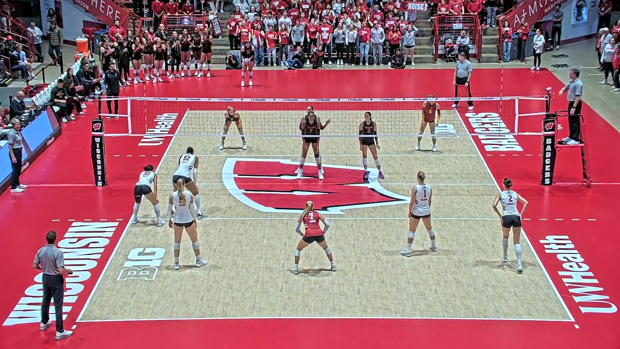 Wisconsin home match filmed from high behind the end line, dead center: net and antennas centered, far end line and both benches in frame, near corners cut off.